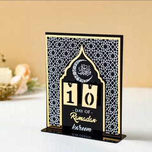 May include: A black and gold Ramadan countdown calendar with a decorative arch design. The calendar features a crescent moon and Arabic calligraphy. The calendar is set to day 10 of Ramadan Kareem.