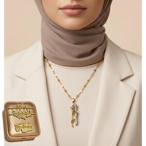 May include: A gold and silver pendant necklace with a chain, featuring two figures embracing. A gold box with the text "SARAH" and "Happy Birthday" is visible. The person is wearing a white blazer and a beige hijab.
