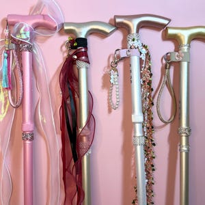 May include: Four walking sticks with T-shaped handles, decorated with ribbons, beads, and rhinestones. The first is pink with a sheer bow, the second is gold with dark red ribbons, the third is white with floral accents, and the fourth is gold.