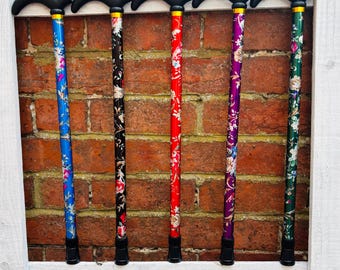 May include: Five floral-patterned walking canes. Each cane has a black handle and tip, with a gold accent. The shafts are blue, black, red, purple, and green, each with a different floral design. The canes are displayed against a brick wall.