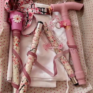 May include: A collapsible mobility cane featuring a pink handle and shaft segments decorated with a cherry blossom pattern. The cane includes a pink velvet ribbon wrap, a white crocheted flower accent, a beaded butterfly charm, and pink rhinestone letter details.