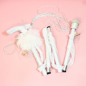 Puede incluir: Un bastón plegable blanco con un mango adornado con plumas blancas, una correa de cuentas y una tapa superior cubierta de pequeñas perlas blancas. El bastón está envuelto en cinta blanca e incluye un dije de plástico transparente con forma de ala.