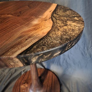 Puede incluir: Una mesa auxiliar redonda hecha a mano con un diseño de borde vivo. La tapa combina madera natural y resina epoxi oscura y brillante. La mesa tiene una sola base de pedestal de madera torneada y un pie redondo, mezclando elementos naturales y modernos. Diámetro: aprox. 60 cm.