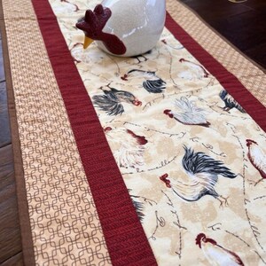 May include: A cream-colored ceramic chicken figurine rests on a decorative table runner. The runner features a repeating rooster pattern with burgundy and tan accents. The runner is approximately 12 inches wide.