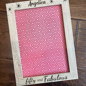 May include: A personalized picture frame with a light wood finish. The frame holds a pink and white geometric patterned print. The top of the frame reads "Angelica" with starburst accents, and the bottom reads "fifty and Fabulous". The frame is 5 x 7 inches.