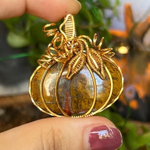 May include: Gold wire wrapped pumpkin pendant with a brown and yellow agate stone. The pendant has a detailed wire design with leaves and a stem.