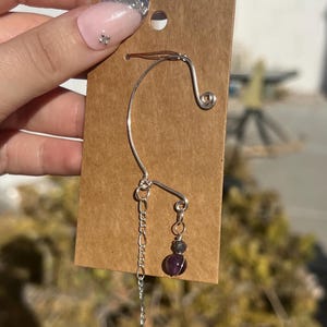May include: A silver ear cuff with a dangling chain and a purple amethyst gemstone. The ear cuff is displayed on a brown card.