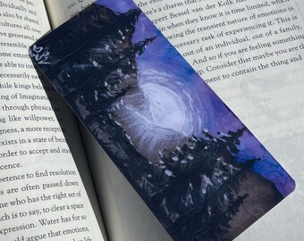 May include: A rectangular tarot card featuring a watercolor painting of a swirling galaxy and dark trees. The card has a purple and blue color scheme. The card is resting on an open book.