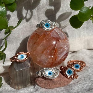 Puede incluir: Una colección de anillos hechos a mano con un diseño de ojo turco. Los anillos están hechos con alambre de plata y cobre, y el ojo turco tiene un diseño azul y blanco. Los anillos se exhiben sobre varias piedras.