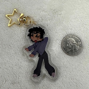 Könnte beinhalten: Ein Acryl-Schlüsselanhänger mit einer Cartoon-Figur mit dunklem, lockigem Haar, die eine violette Jacke und eine dunkle Hose trägt, befestigt an einem goldenen sternförmigen Verschluss. Eine 25-Cent-Münze liegt zum Größenvergleich daneben.