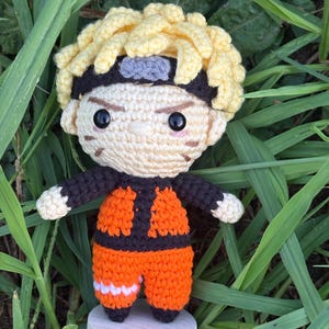 May include: A handmade crochet doll of a character with spiky yellow hair, black headband, and orange outfit with black accents. The doll has black eyes and is standing on a small wooden base. The doll is surrounded by green grass.