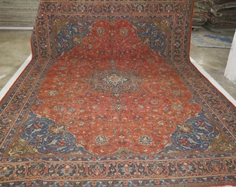 May include: A large, rectangular rug with an intricate floral design. The rug features a predominantly red field with blue and cream-colored accents. The border is a darker blue with floral patterns. The rug is displayed on a white surface.