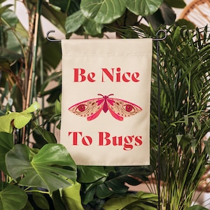 Puede incluir: Bandera de jardín beige con el texto "Be Nice To Bugs" en rojo. Una ilustración de polilla rosa y beige está centrada entre el texto. La bandera se muestra en un jardín con exuberantes plantas verdes.