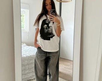 May include: A white t-shirt with a black and white graphic print of a woman. The person wearing the shirt is also wearing dark gray baggy jeans and white sneakers with black accents.