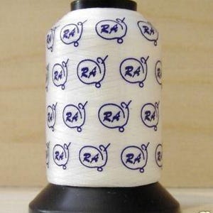 May include: A white spool of thread with a black base and top. The spool is covered in repeating blue circular designs with the letters "RA" inside. The thread is tightly wound, and the spool is set against a light wooden background.