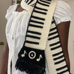 May include: A crochet scarf featuring a piano key design in black and cream. The scarf has a black fringe and musical note accents. The scarf is a unique accessory for music lovers.