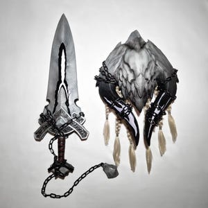 Può includere: Un'esposizione a tema fantasy con una spada d'argento con un design a lama incrinata, avvolta in catene, e un pezzo decorativo abbinato con corna e piume, anch'esso incatenato. Gli oggetti sono esposti su sfondo bianco.