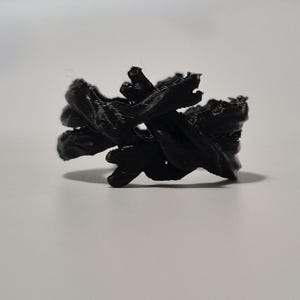May include: A black, 3D printed ring with a unique, abstract design. The ring is made of multiple intertwined strands of plastic, creating a complex and intricate pattern.