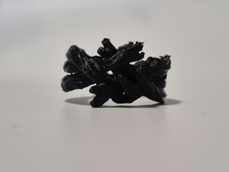 May include: A black, 3D printed ring with a unique, abstract design. The ring is made of multiple intertwined strands of plastic, creating a complex and intricate pattern.