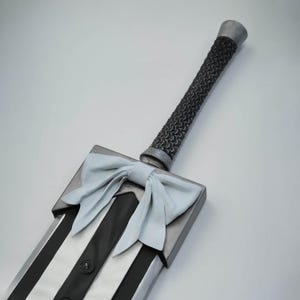 Puede incluir: Una espada blanca y negra con un mango plateado y un lazo blanco atado alrededor del mango.