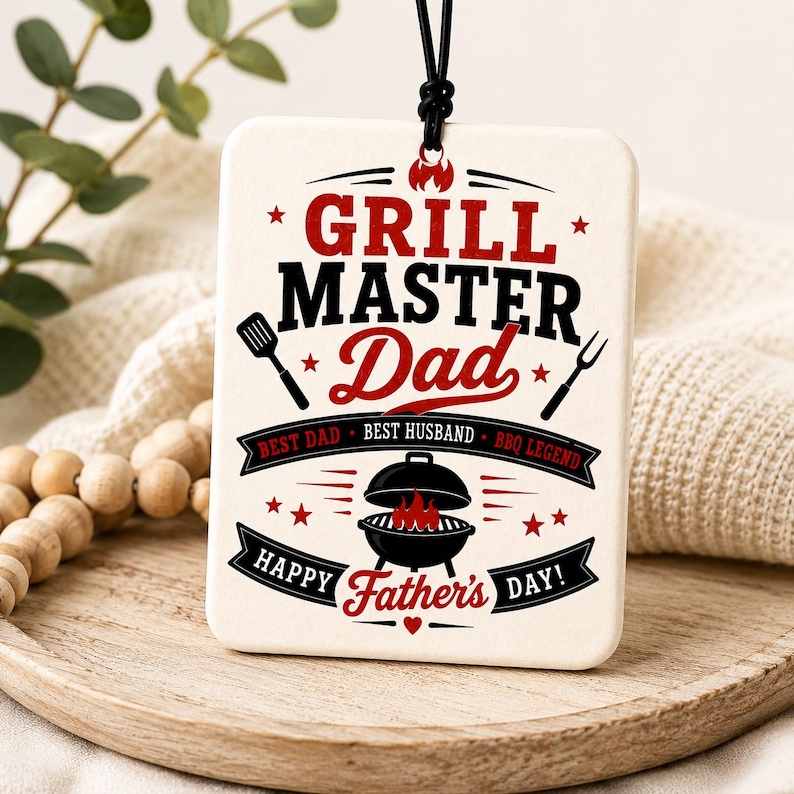 K&ouml;nnte beinhalten: Ein rechteckiges, wei&szlig;es H&auml;ngeornament mit schwarzem und rotem Text, der GRILL MASTER Dad, BEST DAD, BEST HUSBAND, BBQ LEGEND und HAPPY Father's DAY lautet. Das Design enth&auml;lt Abbildungen eines Pfannenwenders, einer Gabel und eines Grills.