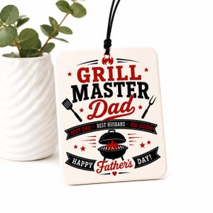 K&ouml;nnte beinhalten: Ein rechteckiges wei&szlig;es H&auml;ngeschild mit schwarzer Kordel und rotem sowie schwarzem Text, der GRILL MASTER Dad, BEST DAD, BEST HUSBAND, BBQ LEGEND und HAPPY Father's DAY! lautet. Das Design enth&auml;lt Abbildungen eines Pfannenwenders, einer Gabel und eines Grills.