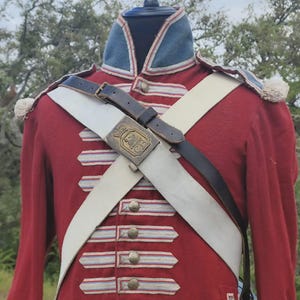May include: A red military jacket with white and gold accents, a white cross strap, and a black belt. The jacket has a blue collar and cuffs with white trim. The jacket is displayed on a black mannequin.