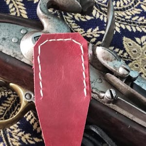 Peut inclure: Un couvre-chien de fusil à silex en cuir rouge avec des coutures contrastantes blanches sur les bords. La protection est placée sur le mécanisme métallique du chien et de la batterie d'une arme à feu en bois avec des détails en laiton.