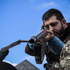 Può includere: Una persona punta un'arma da fuoco d'epoca, con una canna lunga e una lama attaccata. La persona indossa un abito marrone e blu, con un cappello nero. Lo sfondo è un cielo blu. L'arma da fuoco è nera e metallica.