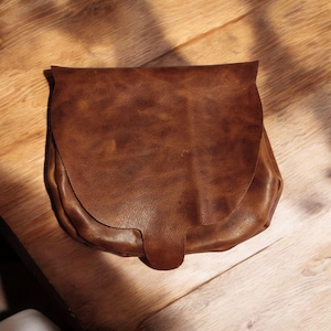 May include: A brown leather satchel bag with a curved flap and a single buckle closure.