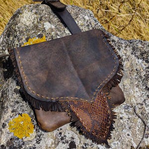 Könnte beinhalten: Eine braune Ledertasche mit Klappe und Fransenbesatz. Die Tasche hat einen Schlangenmuster-Akzent und einen Riemen. Die Tasche liegt auf einem Felsen mit gelbem Flechten.