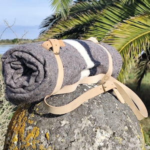 Può includere: Una coperta grigia arrotolata con una striscia bianca, fissata con cinghie in pelle beige. La coperta è appoggiata su una roccia muschiosa, con uno sfondo naturale di vegetazione, acqua e una palma.