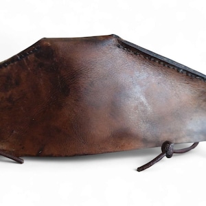 May include: A brown leather arm guard with a curved shape and stitched edges. The leather has a distressed appearance, with variations in colour. It features leather ties for securing it to the arm.