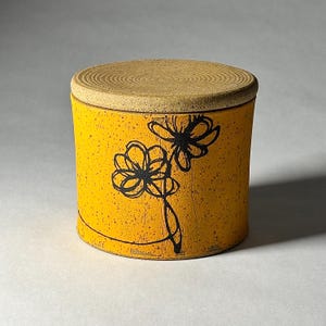 May include: A cylindrical ceramic jar with a lid. The jar is yellow with a speckled texture and features a black floral design. The lid is a light tan color with a ridged texture. The jar is likely used for storage.