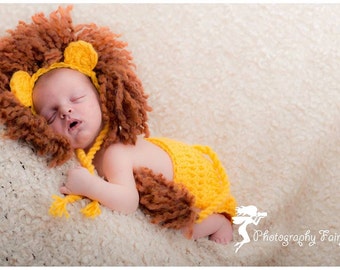 Op de afbeelding: Een pasgeboren baby die een bruin en geel leeuwenkostuum draagt. Het kostuum bestaat uit een wollen muts met oren en een gehaakte staart.