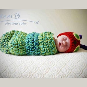 Peut inclure: Un bébé portant un costume de chenille en crochet vert et jaune avec un chapeau de chenille en crochet rouge et jaune. Le bébé dort sur une couverture blanche.