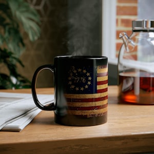 May include: A black ceramic mug featuring a distressed American flag design with the text "1776". The mug has a curved handle and is filled with a hot beverage, with steam rising from the top. The mug is on a wooden surface.