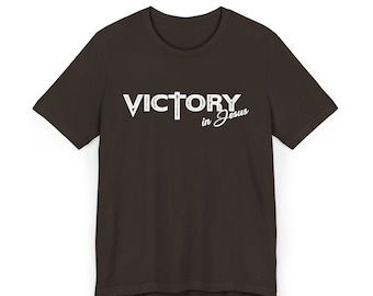 May include: A dark brown t-shirt with the words "VICTORY in Jesus" printed in white. The word "VICTORY" is in a bold, blocky font, with a cross replacing the letter "T". The words "in Jesus" are in a cursive font.