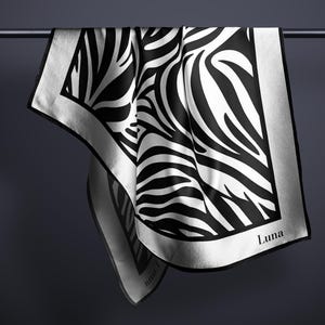 May include: A white silk scarf with a black and white abstract pattern. The scarf has a black border and the word "Luna" is printed in the bottom right corner.