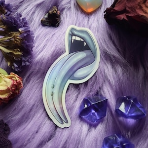 May include: A sticker featuring a stylized mouth with fangs and a long, blue tongue. The sticker has a gradient effect, transitioning from blue to purple. The background is a fluffy, purple surface with crystals and dried flowers.