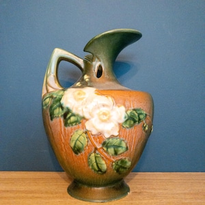 May include: A ceramic pitcher featuring a brown and green gradient glaze with a raised relief of white flowers and green leaves. The vessel has a curved handle and a flared spout, showcasing a textured finish across the rounded body and base.