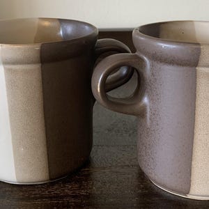 May include: Two ceramic mugs with brown and beige stripes. The mugs have a rounded shape and a handle.