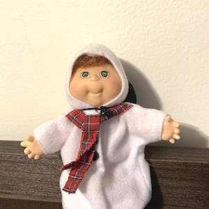 May include: A doll with a white hooded coat and a red and black plaid scarf. The doll has a smiling face with green eyes and brown hair peeking out from under the hood. The doll is a collectible item.