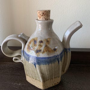 May include: A handmade ceramic oil or vinegar bottle with a cork stopper. The bottle is decorated with brown, blue, and white glaze. The bottle has a unique, twisted handle.