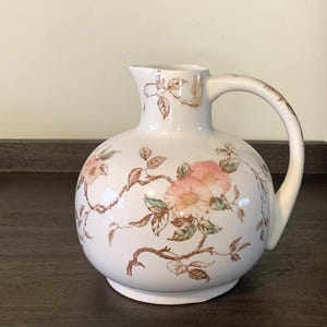 May include: A white ceramic pitcher with a rounded body and a curved handle. The pitcher is decorated with pink and peach-colored floral patterns and brown stems and leaves. The pitcher is sitting on a dark brown surface.