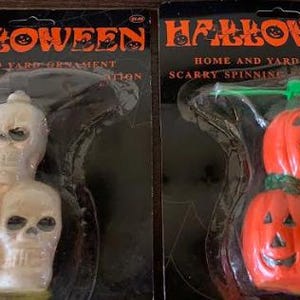 May include: Two Halloween-themed home and yard ornaments. One features a stack of three skulls, and the other has three orange jack-o'-lanterns with green propeller blades. Both are packaged on black backing cards with the word "HALLOWEEN" in orange.