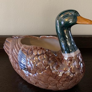 May include: A ceramic duck planter with a brown and green glazed finish. The duck has a realistic design with detailed feathers and a large open beak.