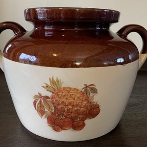 May include: A brown and white ceramic bean pot with two handles. The pot has a pineapple and strawberries design.