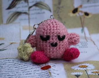 May include: A handmade pink crocheted keychain featuring a round character with closed eyes, small arms, and red feet. A small yellow star is attached to the keychain. The character has a smiling face. The background is a vintage paper.