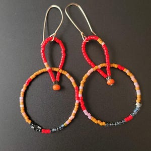 Puede incluir: Un par de pendientes de cuentas hechos a mano. Los pendientes presentan una combinación de colores rojo, naranja, amarillo, rosa y negro. Los pendientes tienen ganchos para las orejas de color plateado y un diseño circular. Los pendientes están hechos de pequeñas cuentas de colores.